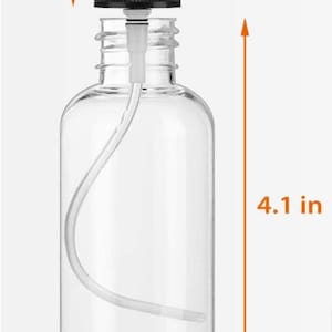 Może przedstawiać: Przezroczysta plastikowa butelka z rozpylaczem z czarną dyszą i przezroczystą nasadką. Butelka ma 10,4 cm wysokości, 3 cm szerokości, a dysza ma 3,6 cm wysokości. Nasadka ma 2,5 cm wysokości. Butelka ma zakrzywioną rurkę.