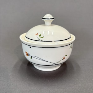 May include: A white ceramic sugar bowl with a lid. The bowl and lid are decorated with black trim and floral designs in orange, green, and black. The bowl has a small foot and a knob on top of the lid.