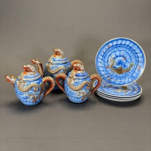 May include: A blue ceramic tea set with dragon motifs. The set includes teapots, a sugar bowl, and plates. The teapots and sugar bowl have dragon-shaped handles and lids. The plates feature a dragon design in the center.