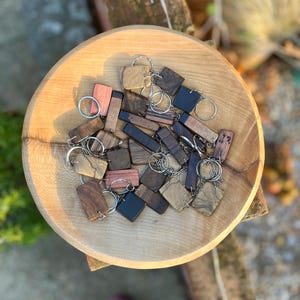 May include: A selection of wooden keyrings in various shapes and wood grains, including rectangular and square designs. The keyrings are displayed in a light brown wooden bowl. The keyrings have silver metal key rings.