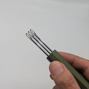 May include: A tool featuring three parallel, silver-colored metal prongs with looped ends, attached to a solid, matte olive-green handle with a hexagonal cross-section. The metal prongs are thin, slender, and have a slight wavy texture along their length.