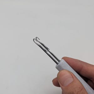 May include: A metal latch hook tool featuring a double-pronged tip and a light gray, speckled handle. The tool is designed with two parallel metal shafts that curve into small hooks at the end, attached to a sturdy, textured gray grip.