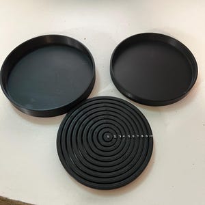 Puede incluir: Tres objetos negros y circulares están dispuestos sobre una superficie blanca. Dos son platos sólidos y poco profundos. El tercero es un disco con un patrón en espiral con los números del 1 al 10 impresos. Los objetos parecen ser de plástico.