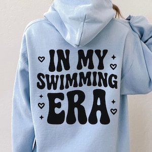 May include: A pale blue hoodie featuring the black text "IN MY SWIMMING ERA", with heart and star motifs. The hoodie has a hood and long sleeves. The design is suitable for casual wear.