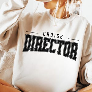 May include: Cream-coloured sweatshirt featuring the text "CRUISE DIRECTOR" in bold, black, block letters. The text is centered on the front of the long-sleeved top. The person wearing the sweatshirt is holding a black mug.
