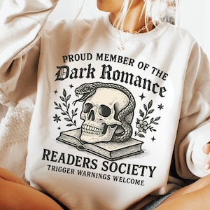 Könnte beinhalten: Cremefarbenes Sweatshirt mit dem Text "PROUD MEMBER OF THE Dark Romance READERS SOCIETY". Die Grafik zeigt einen Totenkopf mit einer Schlange, Blumen und einem offenen Buch. Der Satz "TRIGGER WARNINGS WELCOME" ist ebenfalls aufgedruckt.