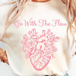 Może przedstawiać: Kremowy t-shirt z różowym anatomicznym sercem i napisem "Go With The Flow". Serce zdobią elementy kwiatowe. Koszulka wykonana jest z miękkiego materiału i nadaje się do noszenia na co dzień.