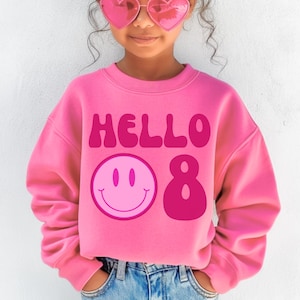 Puede incluir: Una sudadera rosa brillante con el texto "HELLO 8" y una carita sonriente. La niña lleva gafas de sol en forma de corazón y vaqueros azules.