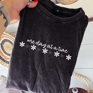 Puede incluir: Camiseta gris oscuro con la frase "one day at a time" en escritura blanca, acentuada con flores blancas y mariposas. La camiseta está doblada y sostenida. El fondo incluye adornos de pared y una imagen enmarcada.