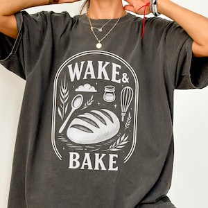 May include: A dark gray oversized t-shirt with a white graphic print that says "Wake & Bake" with a loaf of bread, a spoon, a jar, and a whisk.