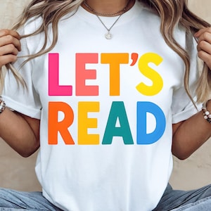 Peut inclure: T-shirt blanc avec l'inscription "LET'S READ" en grosses lettres colorées. Le texte est disposé sur deux lignes, chaque mot étant d'une couleur vive différente. Le t-shirt est de style décontracté.