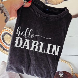 May include: A dark gray t-shirt with the words "hello DARLIN" in white script. The shirt is folded and held up against a white-paneled wall with decorative items in the background. The shirt has a vintage, worn look.