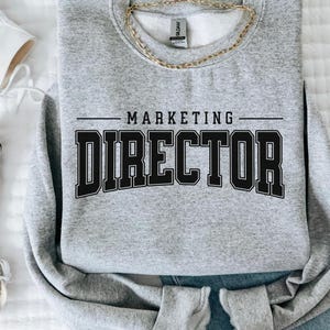 Könnte beinhalten: Ein hellgraues Sweatshirt mit dem Text "MARKETING DIRECTOR" in schwarzen Blockbuchstaben. Eine goldene Halskette und eine blaue Jeans sind ebenfalls im Bild. Weiße Leinenschuhe sind auf der linken Seite des Bildes zu sehen.