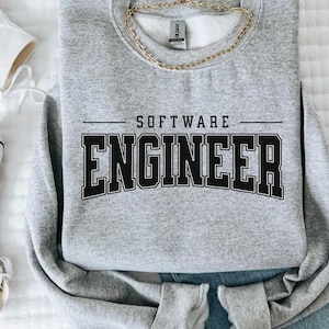 May include: A heather gray sweatshirt with the words "SOFTWARE ENGINEER" printed in bold black letters. A gold chain necklace rests on the sweatshirt. The sweatshirt is paired with blue jeans and white sneakers.