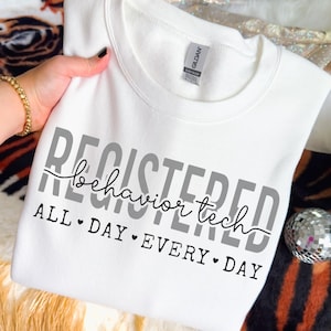 May include: A white crew neck sweatshirt featuring the text "REGISTERED behavior tech ALL DAY EVERY DAY" in grey and black. The sweatshirt is made of a soft material, suitable for casual wear.