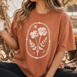 May include: A rust-colored short-sleeve t-shirt featuring a white graphic print of two flowers and leaves enclosed within an oval frame with small star accents. The shirt has a crew neckline and a relaxed fit, paired with solid black leggings.