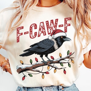 May include: Cream-colored t-shirt featuring a black crow wearing a Santa hat, perched on a branch with Christmas lights. The text "F-CAW-F" is printed above the crow in red letters. A festive holiday design.