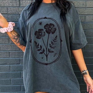 May include: Dark gray oversized t-shirt featuring a floral design within an oval frame. The graphic includes two flowers, leaves, and stars. The shirt has short sleeves and a relaxed fit, suitable for casual wear.
