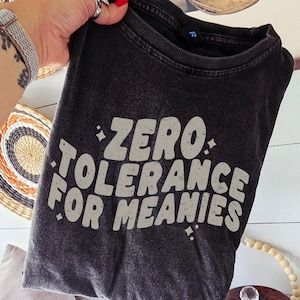 Op de afbeelding: Donkergrijs t-shirt met de witte tekst "ZERO TOLERANCE FOR MEANIES" in gestileerde letters. Het shirt wordt door een hand omhoog gehouden, met een geweven wandkleed en een ingelijste foto op de achtergrond.