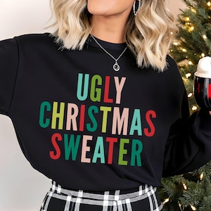 Może przedstawiać: Czarna bluza z długim rękawem z napisem UGLY CHRISTMAS SWEATER wydrukowanym wielokolorową czcionką w odcieniach czerwieni, zieleni i jasnego różu. Ubiór zestawiono z czarno-białą spódnicą w kratę, co tworzy świąteczny wygląd.