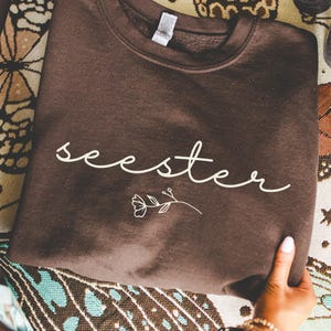 以下が含まれることがあります： 筆記体で「seester」の文字と小さな花の模様が入った茶色のスウェットシャツ。 スウェットシャツは折りたたまれ、手で持たれており、背景には模様のある布があります。