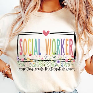 Peut inclure: T-shirt crème avec l'inscription "SOCIAL WORKER" en arc-en-ciel dans un rectangle noir, surmonté d'un cœur rose. En dessous, un motif floral et les mots "planting seeds that last forever".