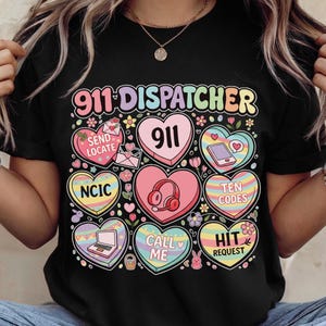 May include: Black t-shirt with the text "911 DISPATCHER" in rainbow colors. The shirt features heart-shaped graphics with phrases like "SEND LOCATE", "911", "NCIC", "TEN CODES", "CALL ME", and "HIT REQUEST".
