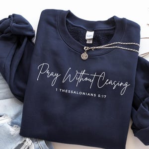 May include: Navy blue sweatshirt with the text "Pray Without Ceasing" and the scripture reference 1 Thessalonians 5:17 in white script. A silver necklace with a pendant rests on the sweatshirt. The sweatshirt is folded next to a pair of distressed denim jeans.
