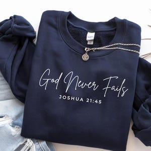 May include: Navy blue sweatshirt with the text "God Never Fails" and "Joshua 21:45" in white script. A silver necklace with a pendant rests on the sweatshirt. The sweatshirt is paired with distressed denim jeans.