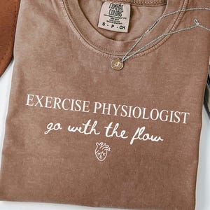 Può includere: Una maglietta marrone con le parole "EXERCISE PHYSIOLOGIST go with the flow" stampate in bianco. Una collana d'argento con un piccolo ciondolo è drappeggiata sulla maglietta. La maglietta ha un girocollo e maniche corte.