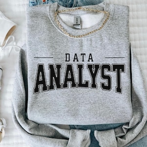 May include: A heather gray sweatshirt with the words "DATA ANALYST" in bold, black, collegiate-style lettering. A gold chain necklace is draped across the top. The sweatshirt is folded on top of blue jeans and white sneakers.