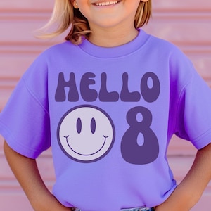 Pode incluir: Uma camisola lilás de manga curta com as palavras "HELLO 8" em roxo escuro. Um rosto sorridente branco e o número 8 estão abaixo do texto. A camisola é usada por uma pessoa com óculos de sol redondos.