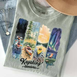 May include: A light green t-shirt with a watercolor design of four different kayaking scenes. The scenes include a starry night, a forest, a sunset, and a lake with a aurora borealis. The text "Kayaking Adventures" is printed below the scenes.