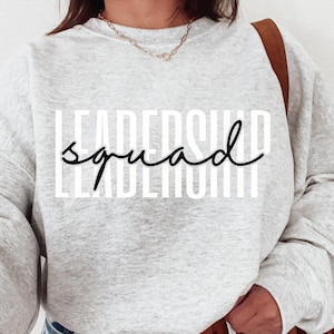 May include: A light gray sweatshirt with the words "LEADERSHIP squad" printed in white and black. The sweatshirt has long sleeves and is paired with blue jeans and a brown belt. A brown shoulder bag is visible.