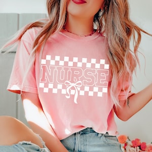 Op de afbeelding: Een koraalkleurig T-shirt met korte mouwen en een witte grafische print op de borst. Het ontwerp bestaat uit een geblokte rechthoekige achtergrond met het woord NURSE in dikke blokletters en een klein strikicoontje onder de tekst.
