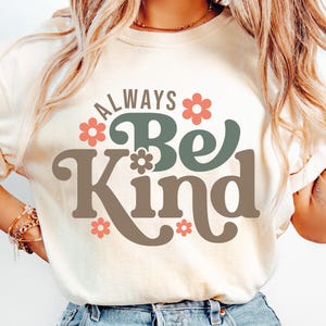 May include: A cream-coloured t-shirt featuring the phrase "ALWAYS Be Kind" in a retro font, with coral and teal floral accents. This casual wear item promotes a positive message and is suitable for everyday wear.