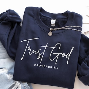 Puede incluir: Sudadera azul marino con la frase "Trust God" en escritura blanca. Debajo está escrito "Proverbs 3:5". Una cadena plateada con un colgante está colocada sobre la sudadera.