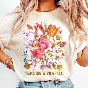 May include: A cream-coloured t-shirt featuring a vibrant watercolour-style floral bouquet with tulips, peonies, and small blossoms in shades of pink, red, orange, and purple. The text "TEACHING WITH GRACE" is printed in brown letters below the flowers.