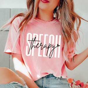 Könnte beinhalten: Ein korallenfarbenes kurzärmeliges T-Shirt mit dem Wort SPEECH in großen weißen Blockbuchstaben und dem Wort Therapy in schwarzer Schreibschrift mit einem kleinen Herzsymbol am Ende, auf die Brust gedruckt.