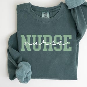 Może przedstawiać: Teal bluza z napisem "NURSE" dużymi, jasnozielonymi literami blokowymi. Słowo "nurse" jest napisane kursywą nad literami blokowymi. Rękawy są podwinięte.