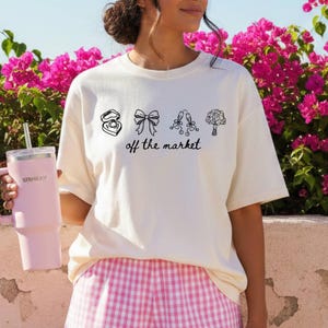 May include: A cream-coloured t-shirt featuring black line art of a ring box, bow, champagne flutes, and a bouquet, with the text "off the market". The person is wearing pink and white checked shorts and holding a pink tumbler.