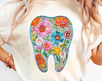 May include: Cream-colored t-shirt featuring a large, embroidered tooth design. The tooth is light blue and filled with colorful flowers in shades of pink, orange, yellow, and blue. The design is centered on the front of the shirt.