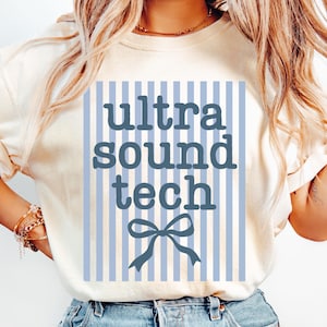 May include: Cream-colored t-shirt with the text "ultra sound tech" in a blue font, set against a blue and white striped background. A blue bow is below the text. The shirt is made of a soft material.