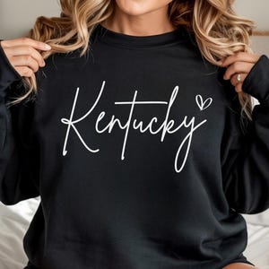 May include: Black sweatshirt featuring the word "Kentucky" in white cursive script, with a small heart symbol. The garment is a plain black colour.