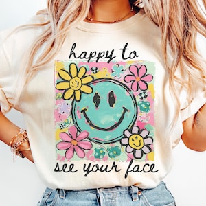 May include: Cream-coloured t-shirt with a colourful graphic featuring a turquoise smiley face surrounded by flowers. The text "happy to see your face" is above and below the graphic. The shirt has short sleeves.