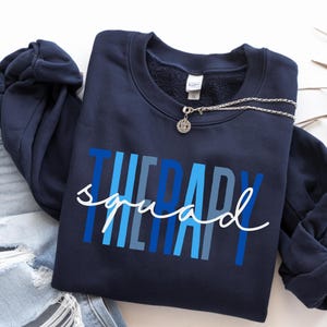 Op de afbeelding: Marineblauw sweatshirt met de tekst "THERAPY squad" in blauwe en witte tinten. Een zilveren ketting met een ronde hanger ligt op het sweatshirt. Het sweatshirt wordt gecombineerd met een distressed jeans.