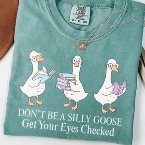 May include: A teal Comfort Colors t-shirt featuring three cartoon geese wearing glasses and holding books. The text "DON'T BE A SILLY GOOSE Get Your Eyes Checked" is printed below the geese. A gold necklace with a pendant is draped on the shirt.