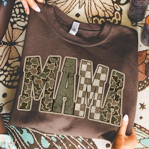 May include: A brown sweatshirt with the word "MAMA" in a camouflage and checkered design. The sweatshirt is folded and held by a person. The background includes candles and a decorative cloth.