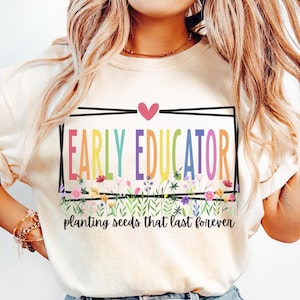 May include: Cream-colored t-shirt with the words "EARLY EDUCATOR" in colorful letters inside a black rectangle. Below the text is a floral design with the words "planting seeds that last forever". A small pink heart is above the text.