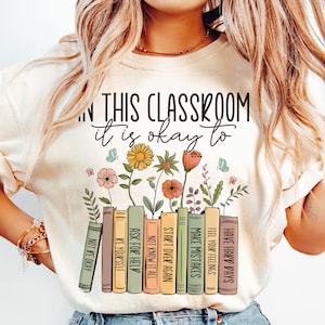 May include: A cream-coloured t-shirt featuring the text "IN THIS CLASSROOM it is okay to" above a row of books with floral designs. The books display phrases such as "Make Mistakes" and "Have Hard Days".